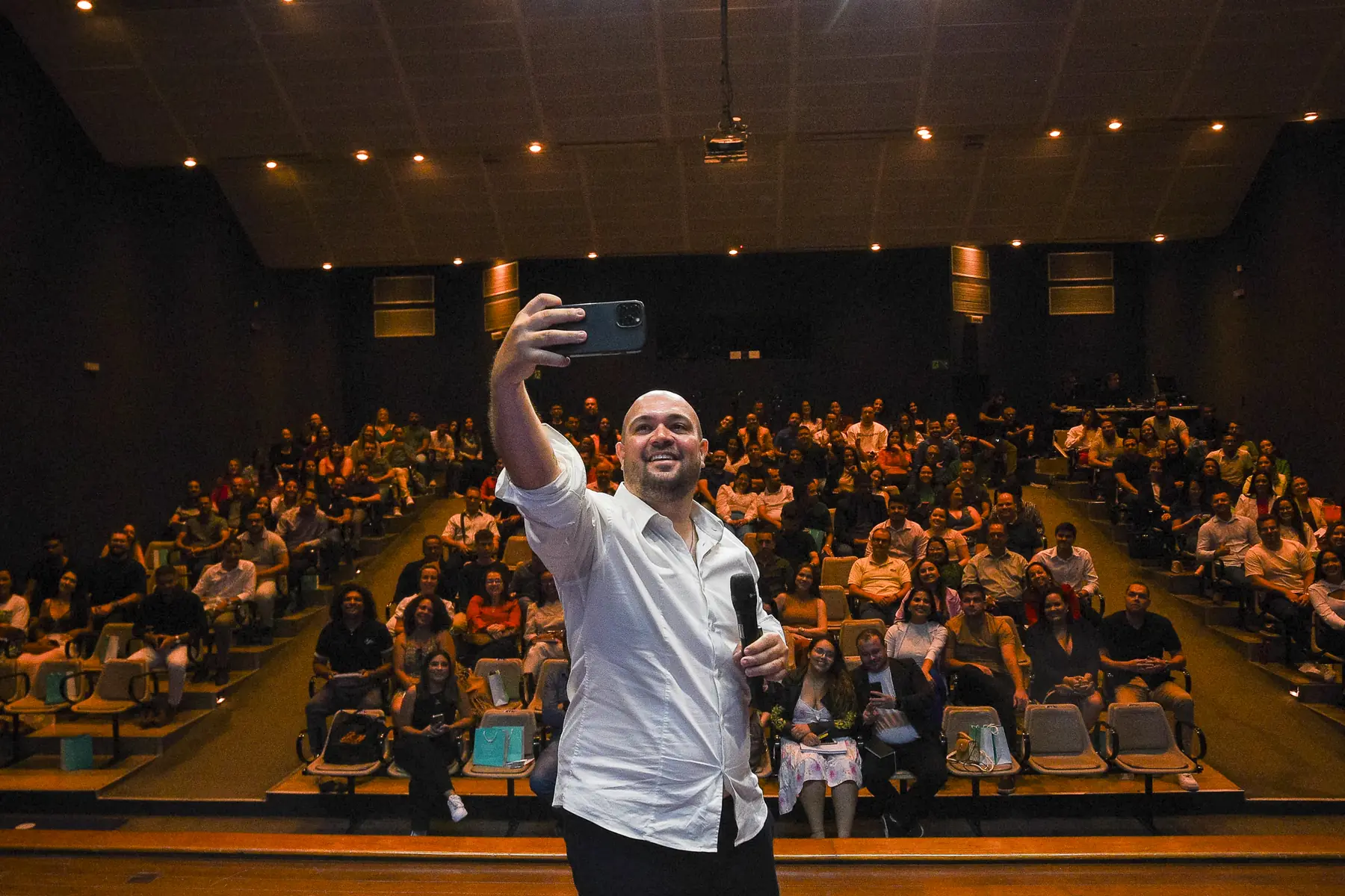 Alex Vilela no palco tirando uma selfie com o auditório cheio ao fundo